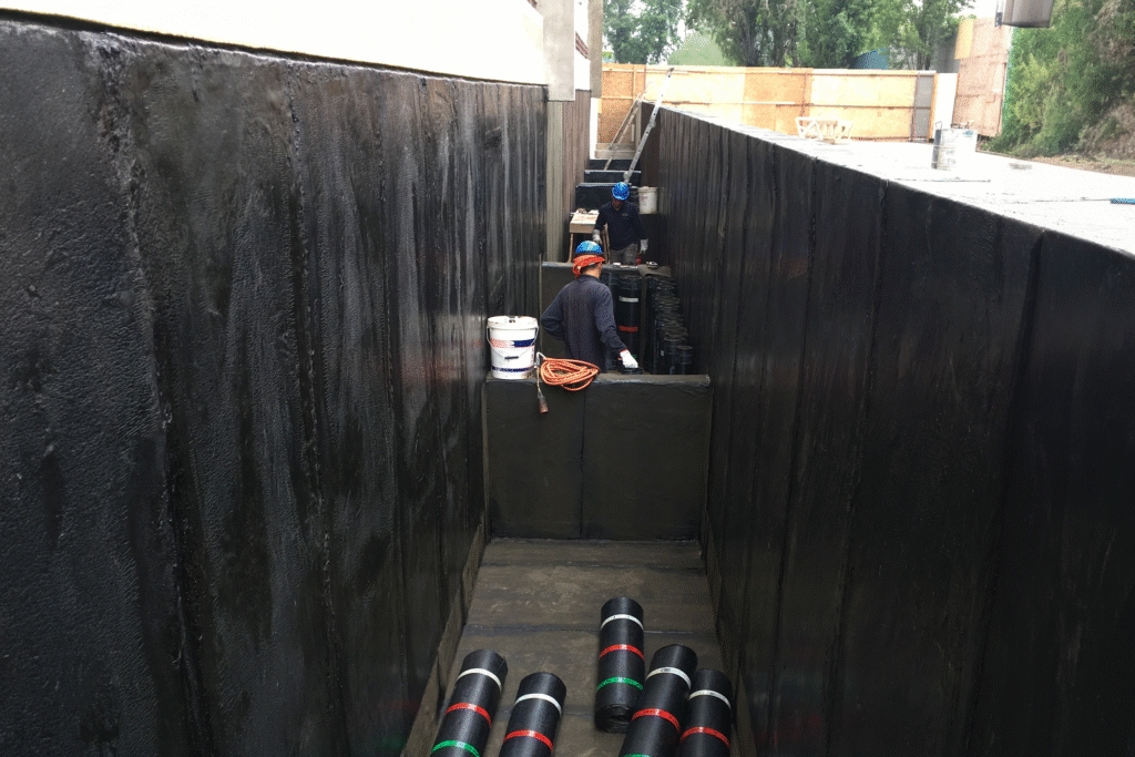 Trabajadores aplicando membrana asfáltica para impermeabilizar muros de contención y fundaciones