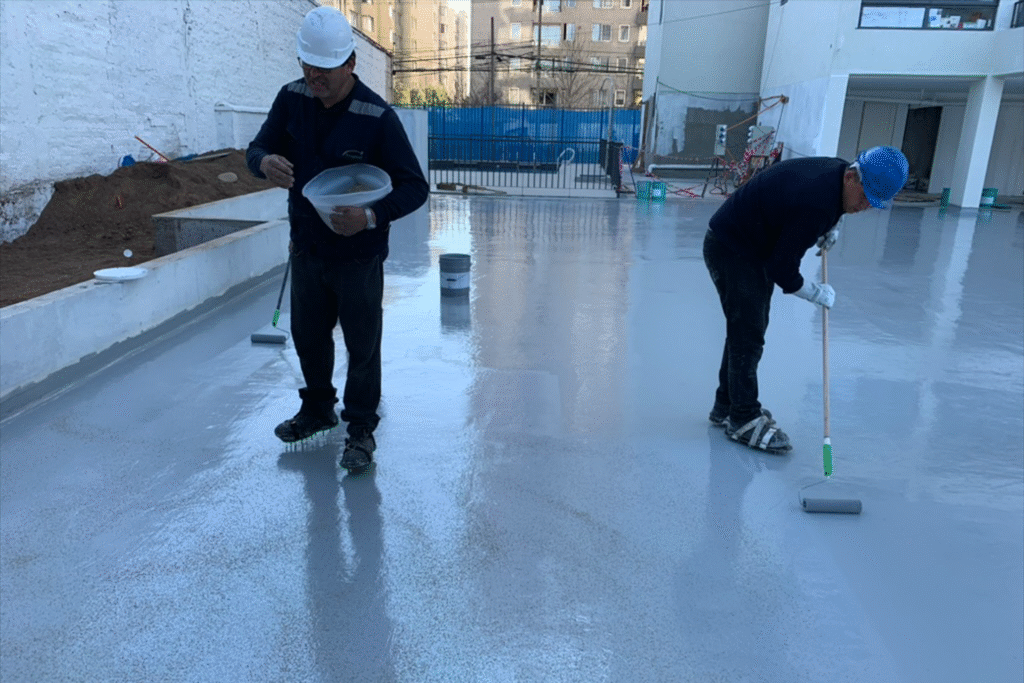 Trabajadores aplicando recubrimiento impermeabilizante en piso de hormigón para estacionamiento con rodillos.