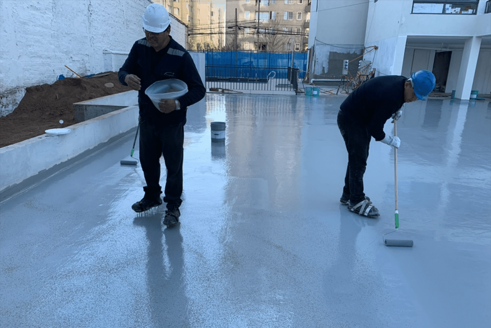 Trabajadores aplicando recubrimiento impermeabilizante en piso de hormigón para estacionamiento con rodillos.