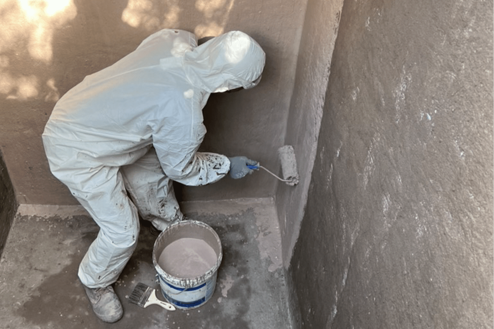 Trabajador aplicando pintura impermeabilizante en estanque de agua con rodillo y traje de protección.